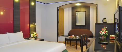 A hotel room with a large bed, a desk with a chair, a small table with a cup and flowers, and a TV on a stand.