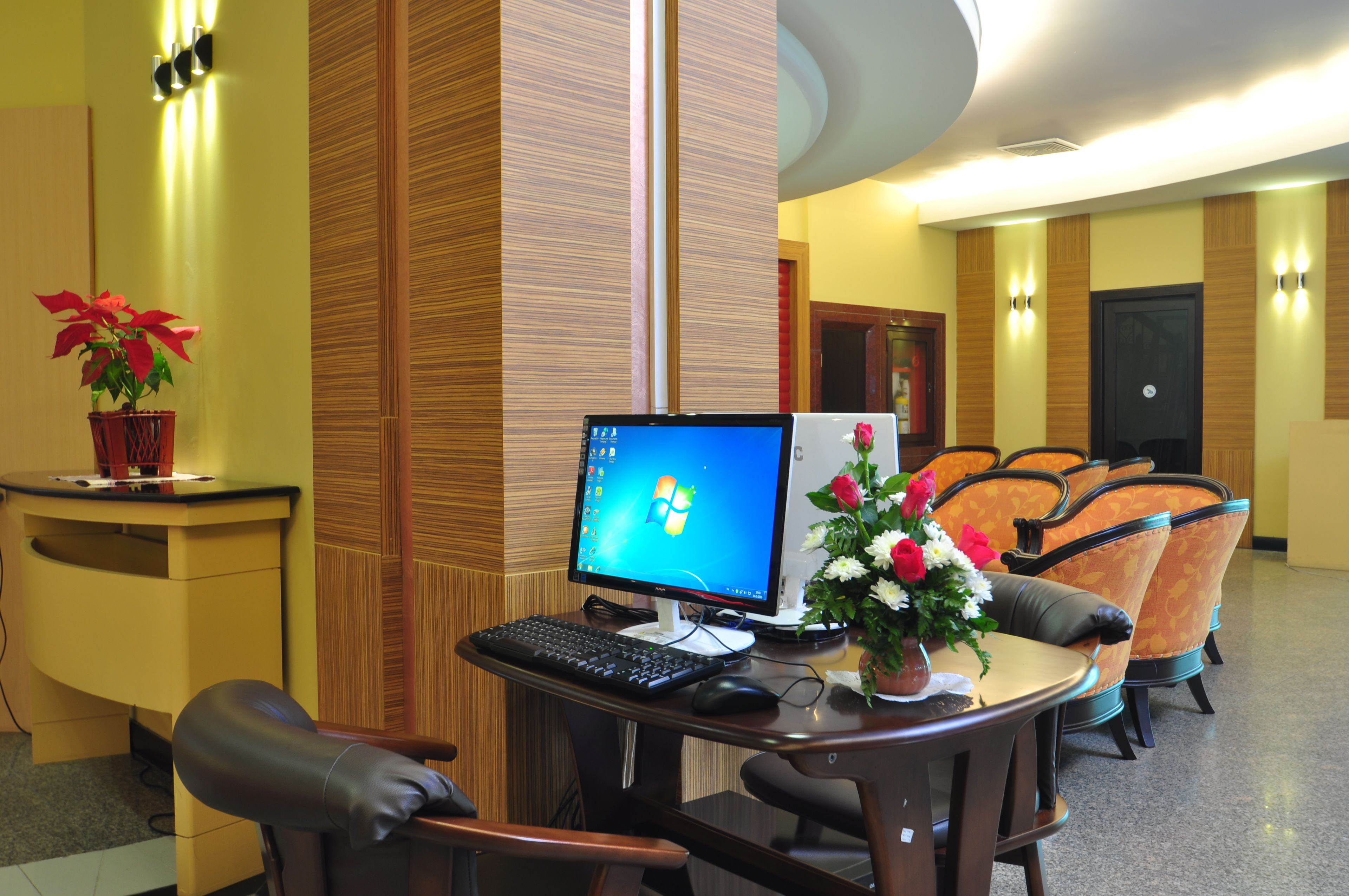 Un vestíbulo de hotel con una recepción, un escritorio con monitor, teclado y ratón, y un área de descanso con sillas.