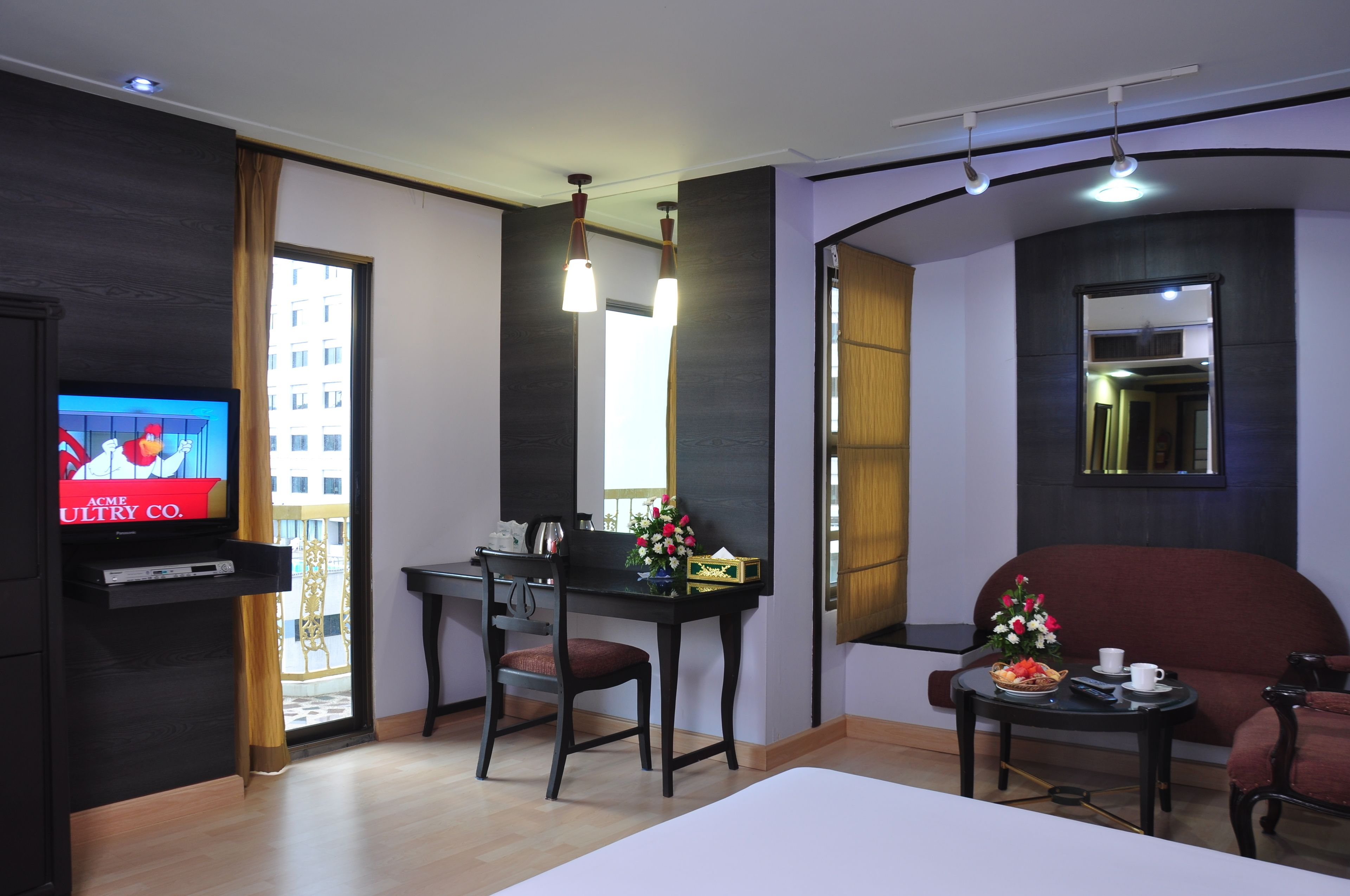 A modern hotel room with a TV, a dining area, a sofa, and a small table with flowers.