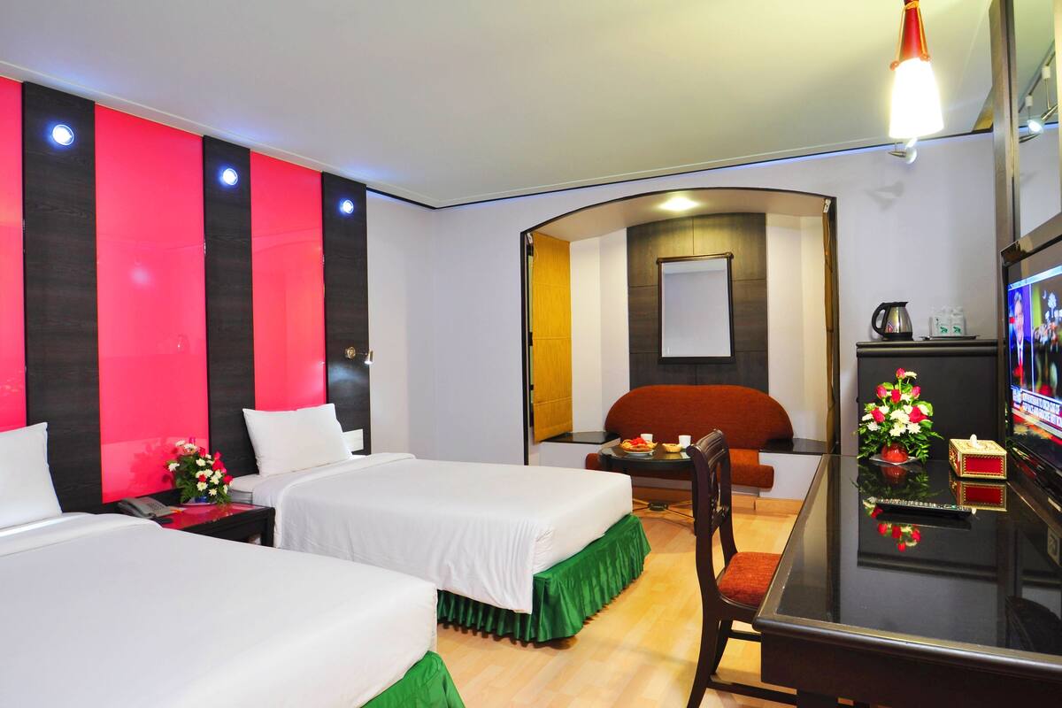 A hotel room with two beds, a desk, a chair, a TV, and a small table with a flower vase.