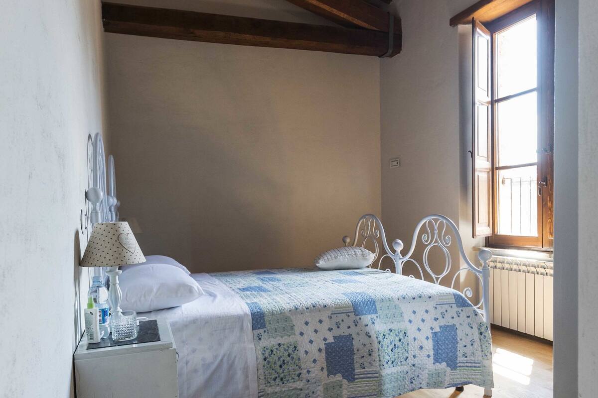 Ein Schlafzimmer mit Holzboden, einem Bett mit gemustertem Überwurf, einem Nachttisch mit Lampe und einem Fenster mit Blick.