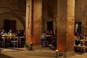 Un área de comedor al aire libre con columnas y arcos de ladrillo, mesas con sillas y algunas personas cenando.