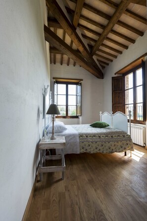 Un dormitorio con techo de madera, una cama con colcha estampada, un cojín verde, una mesita de noche con lámpara y una ventana con persianas de madera.