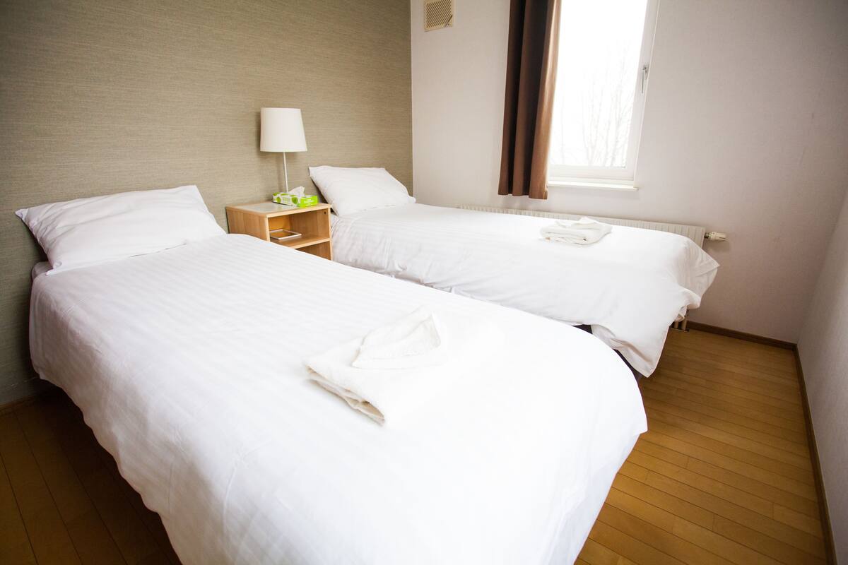 Hotellihuone, jossa on kaksi erillistä sänkyä, yöpöytä lampun kanssa ja ikkuna, jossa on verhot.