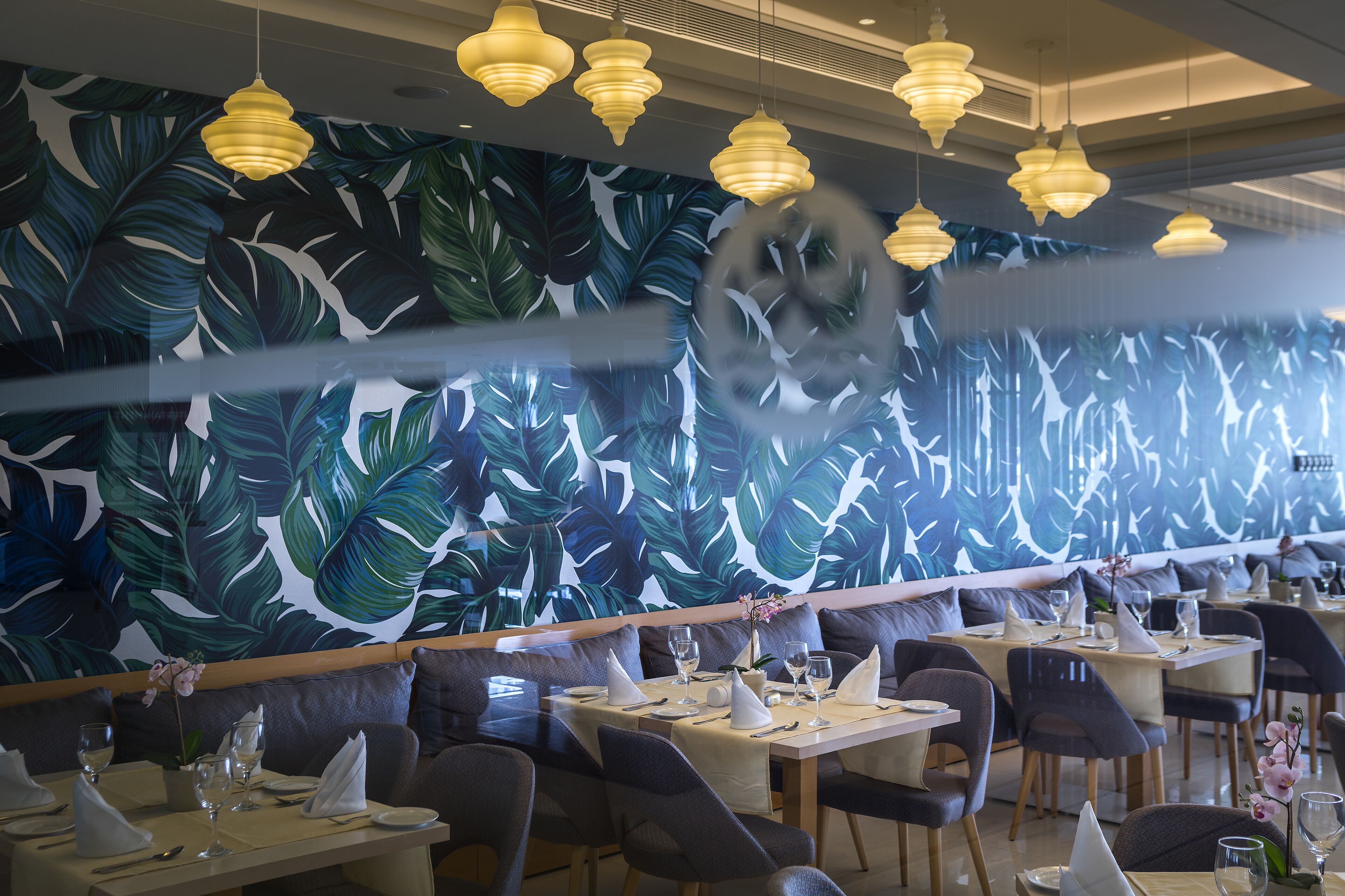 En restaurang med en väggmålning som föreställer tropiska blad, flera matbord och hängande lampor.