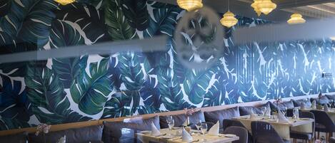 Un restaurante con un mural de hojas tropicales, varias mesas de comedor e iluminación colgante.