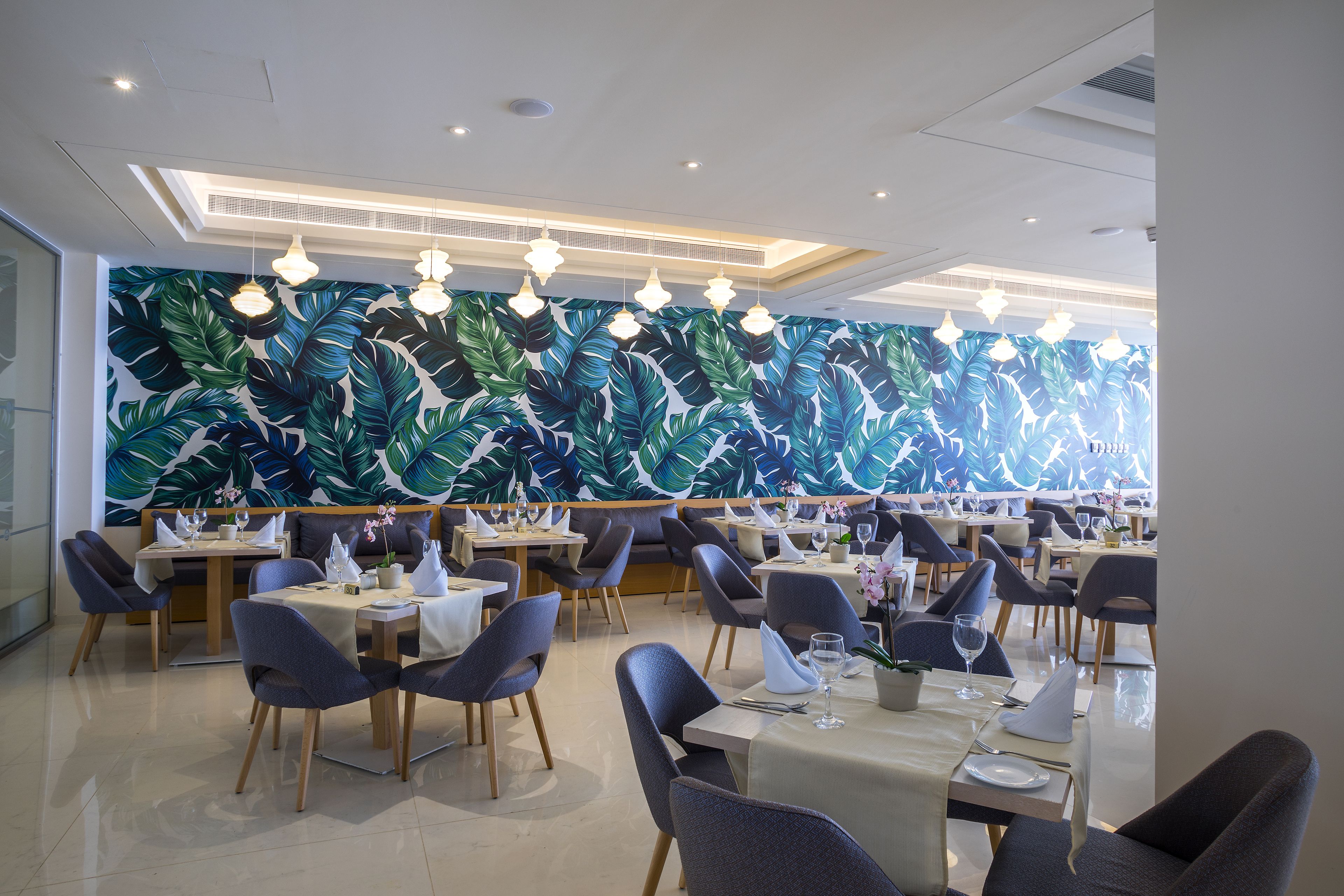 A restaurant with tables and chairs, a large tropical leaf mural on the wall, and hanging pendant lights.