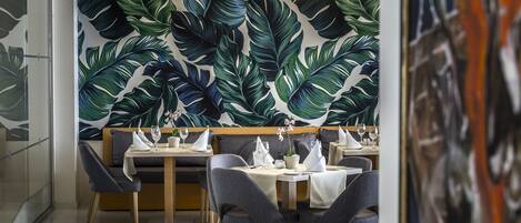 A modern restaurant interior with a tropical leaf wallpaper, wooden tables, and grey upholstered chairs.