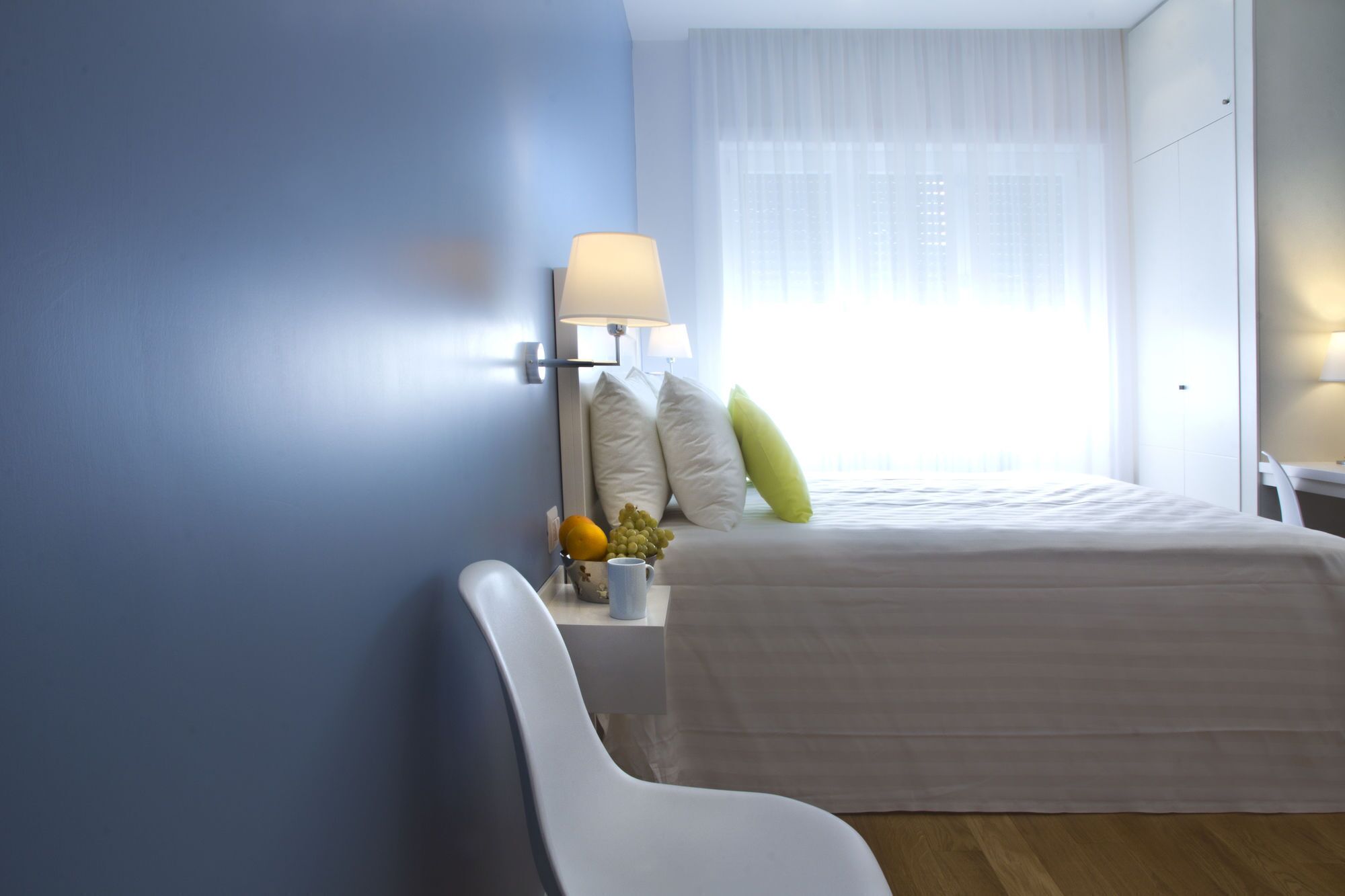 Ein modernes Hotelzimmer mit einem Bett, einem Nachttisch mit Lampe, einem Sessel und einem Fenster mit durchsichtigen Vorhängen.