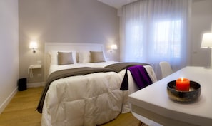 Et hotelværelse med en seng, sengeborde med lamper, et skrivebord med et lys og et vindue med gardiner.
