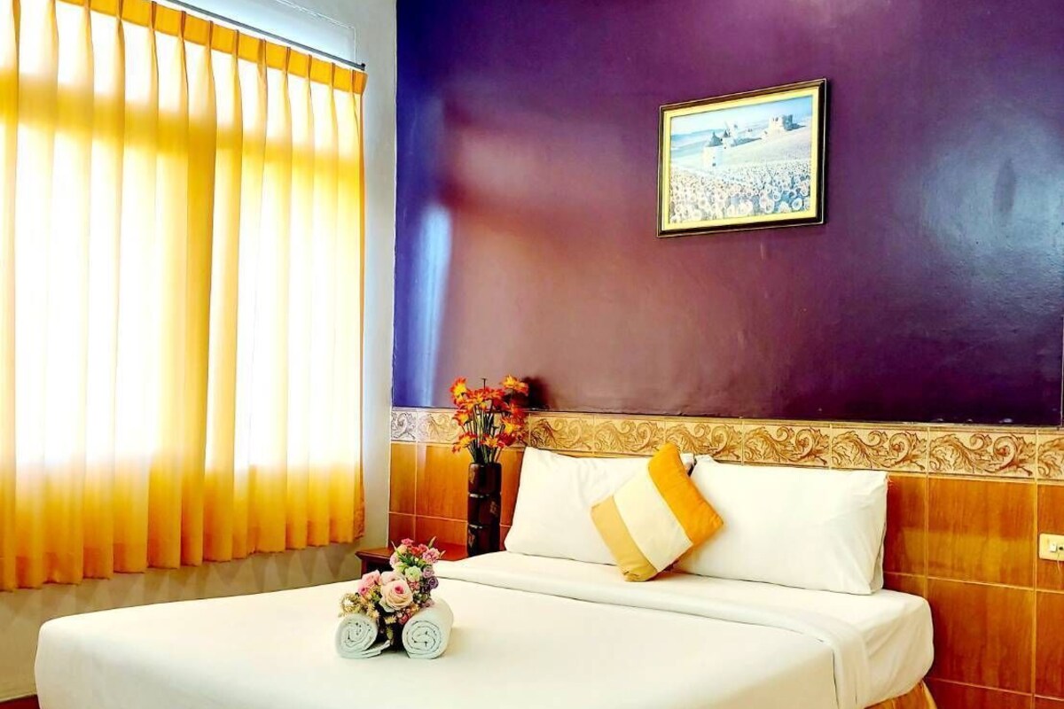 Una camera d'albergo con un letto, tende, una ventola a soffitto, un quadro appeso al muro e un vaso con fiori.