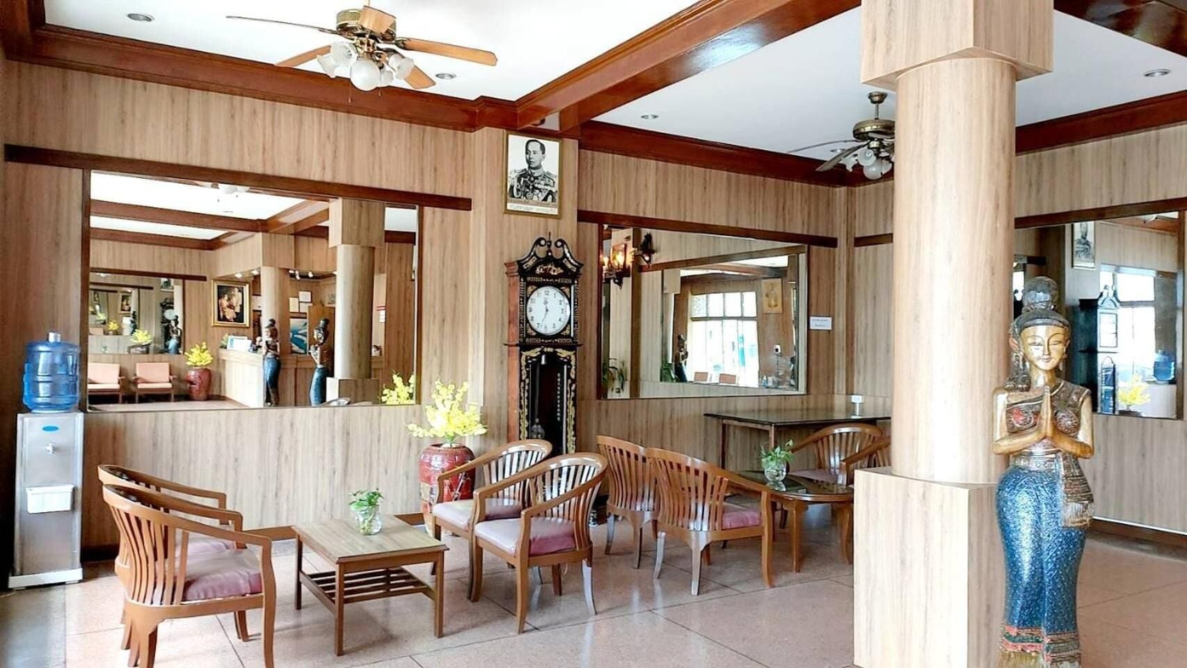 A spacious hotel lobby with wooden paneling, a central seating area with chairs and a table, a water dispenser, a clock, and a statue.