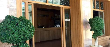 A building entrance with a sign reading "TISUDISUABANBUYS" and a reception desk inside. There are two potted trees in front.
