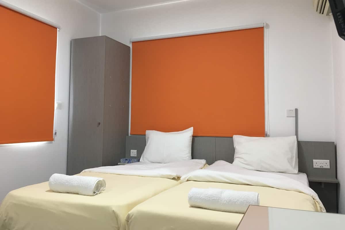 A hotel room with two beds, orange window shades, a grey wardrobe, and a white nightstand.