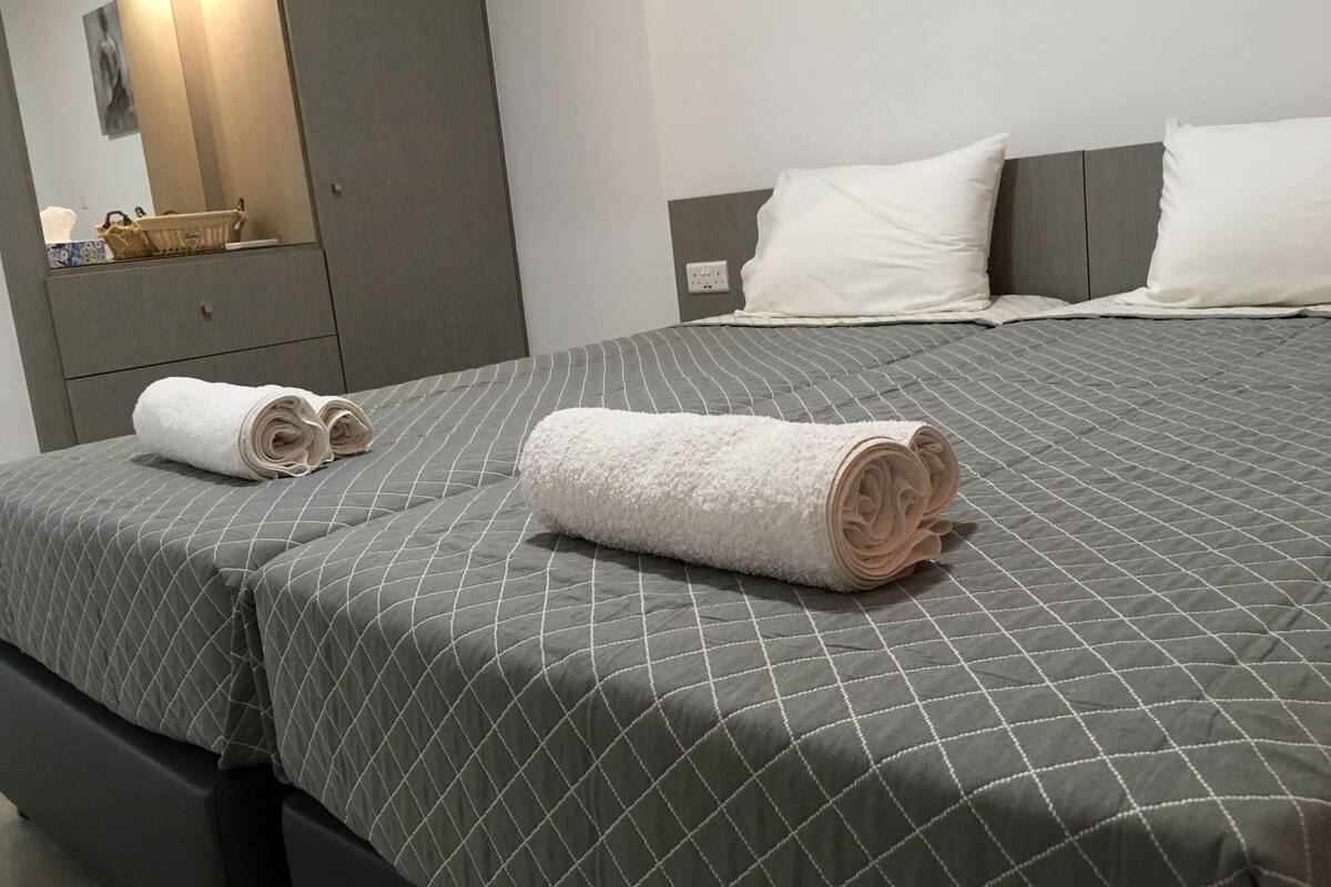 A neatly made bed with a grey quilted bedspread and two rolled white towels on the side.
