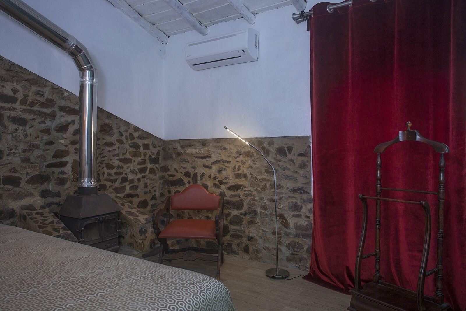 Een kamer met een stenen muur, een fluwelen gordijn in rood, een houten stoel, een bed met beddengoed met een patroon, en een houtkachel.