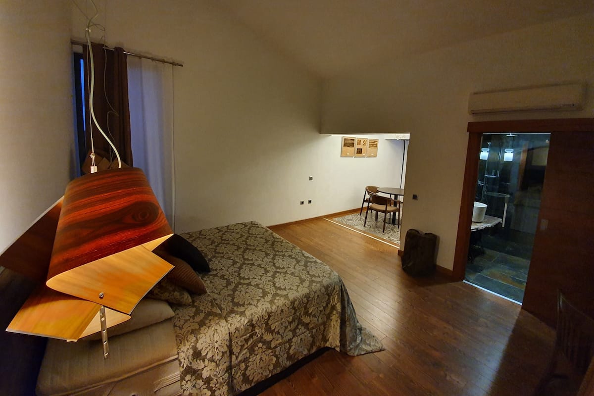 A bedroom with a bed, a desk, and a chair. There is a window with curtains, a door, and a wall-mounted shelf with pictures.