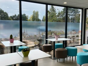 Un espace de restauration moderne, avec des tables et des chaises, de grandes fenêtres offrant une vue sur le paysage urbain, et des plantes d’ornement en pot.
