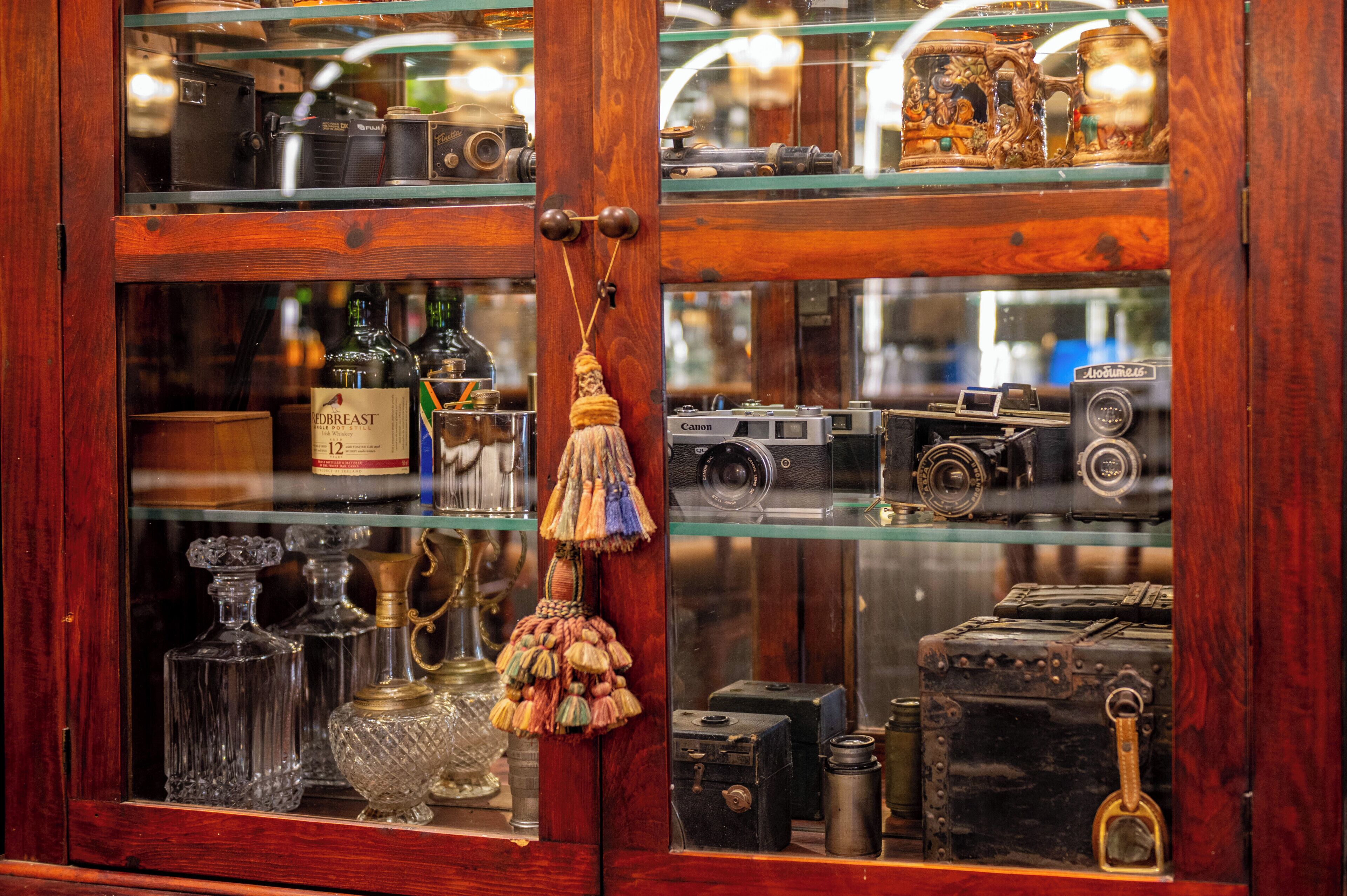 Een houten kast met glazen deuren die vintage camera's, flessen en een messing lamp toont.