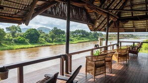 Terraza de madera con mobiliario de mimbre que da a un río y vegetación.