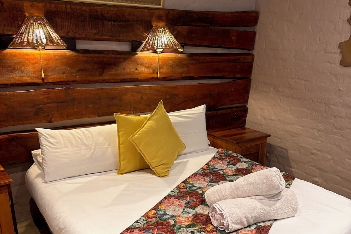 A neatly made bed with white and yellow pillows, a floral bedspread, and a folded towel on the side. There are two bedside lamps on wooden nightstands, and a framed picture hangs on the wall above the bed.
