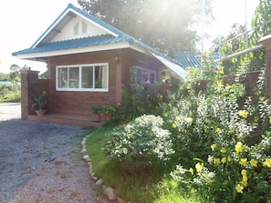 A small house with a blue roof, a brick exterior, and a front porch. There are potted plants and flowering bushes in the front yard. - I Din Lake View Resort Nakhon Nayok (Nakhon Nayok)