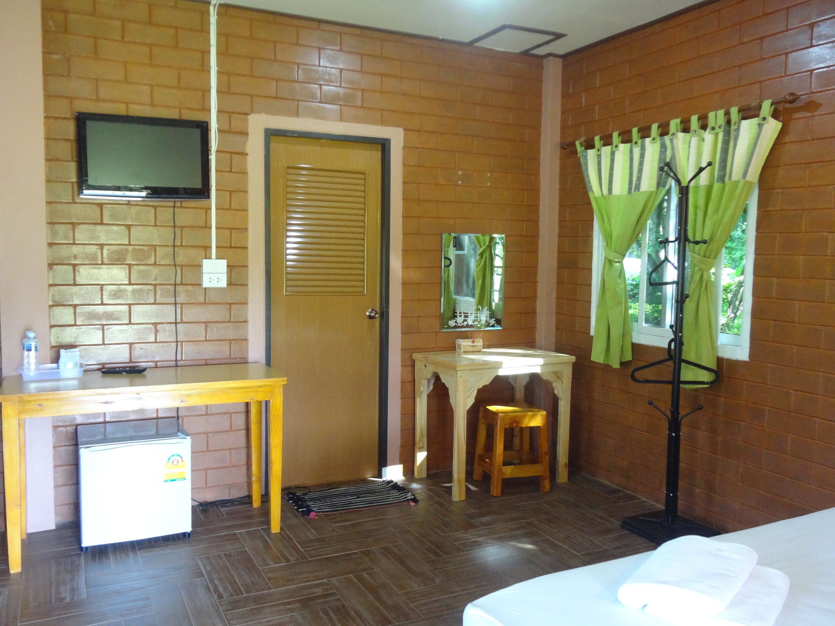 Ein Zimmer mit Backsteinwand, einem Holztisch mit Fernseher, einem kleinen Kühlschrank, einem hölzernen Hocker, einem Spiegel und einem Fenster mit grünen Vorhängen.