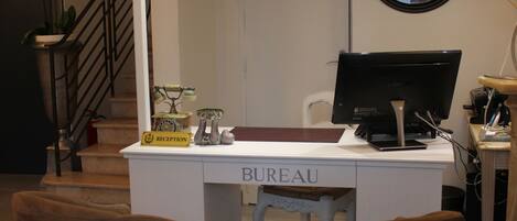 Un comptoir d’accueil avec une enseigne indiquant « RÉCEPTION » et « BUREAU », deux chaises marron devant, un miroir rond accroché au mur et un escalier en arrière-plan.