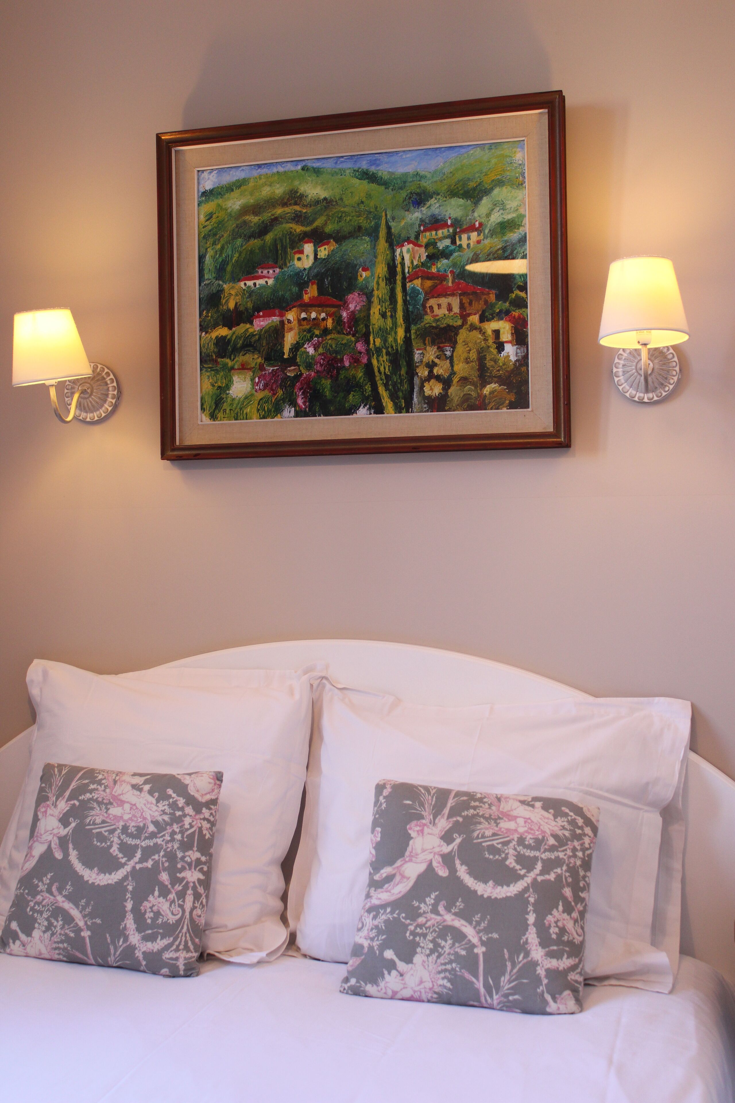 A hotel room with a bed, two decorative pillows, a framed painting on the wall, and two wall-mounted lamps.
