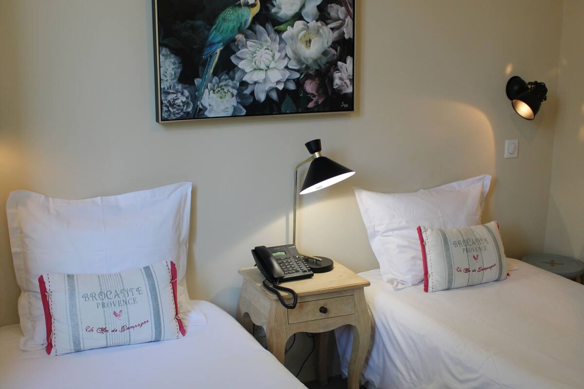 Une chambre d’hôtel avec deux lits, une table de chevet avec une lampe, un tableau floral encadré au mur et un coussin orné d’un motif.