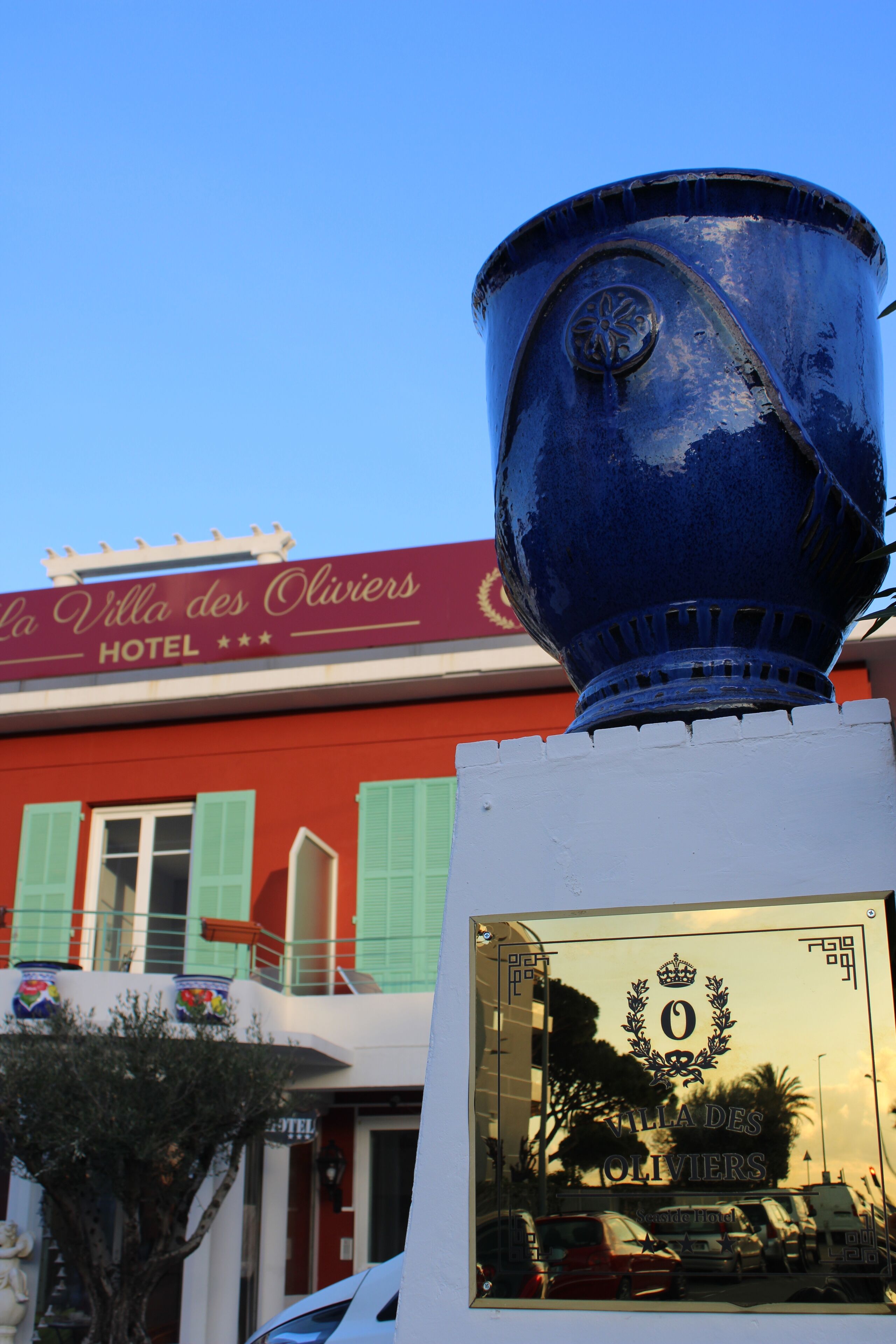 Un trofeo azul sobre un pedestal blanco, con el reflejo del letrero de Villa des Olivier y una palmera.