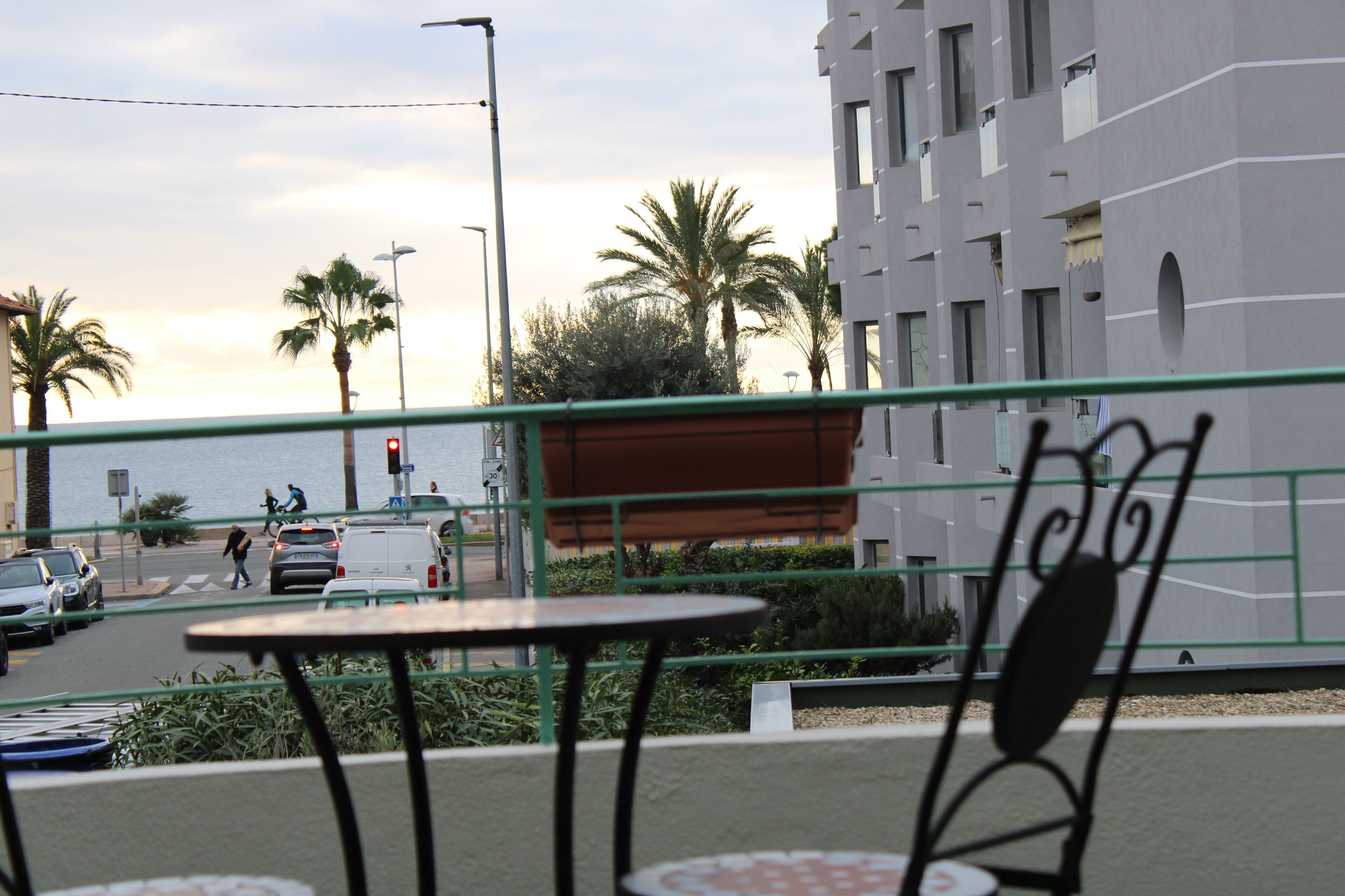 A view from a balcony overlooking a street with palm trees, a body of water, and a building with balconies. There are tables and chairs on the balcony.