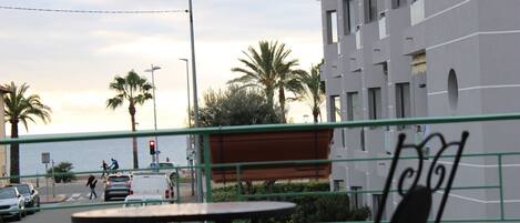 A view from a balcony overlooking a street with palm trees, a body of water, and a building with balconies. There are tables and chairs on the balcony.