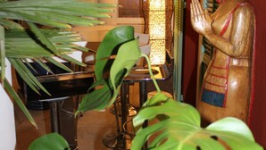 Een beeld van een vrouw in traditionele kledij, een piano en weelderige groene planten in een warm verlichte ruimte.