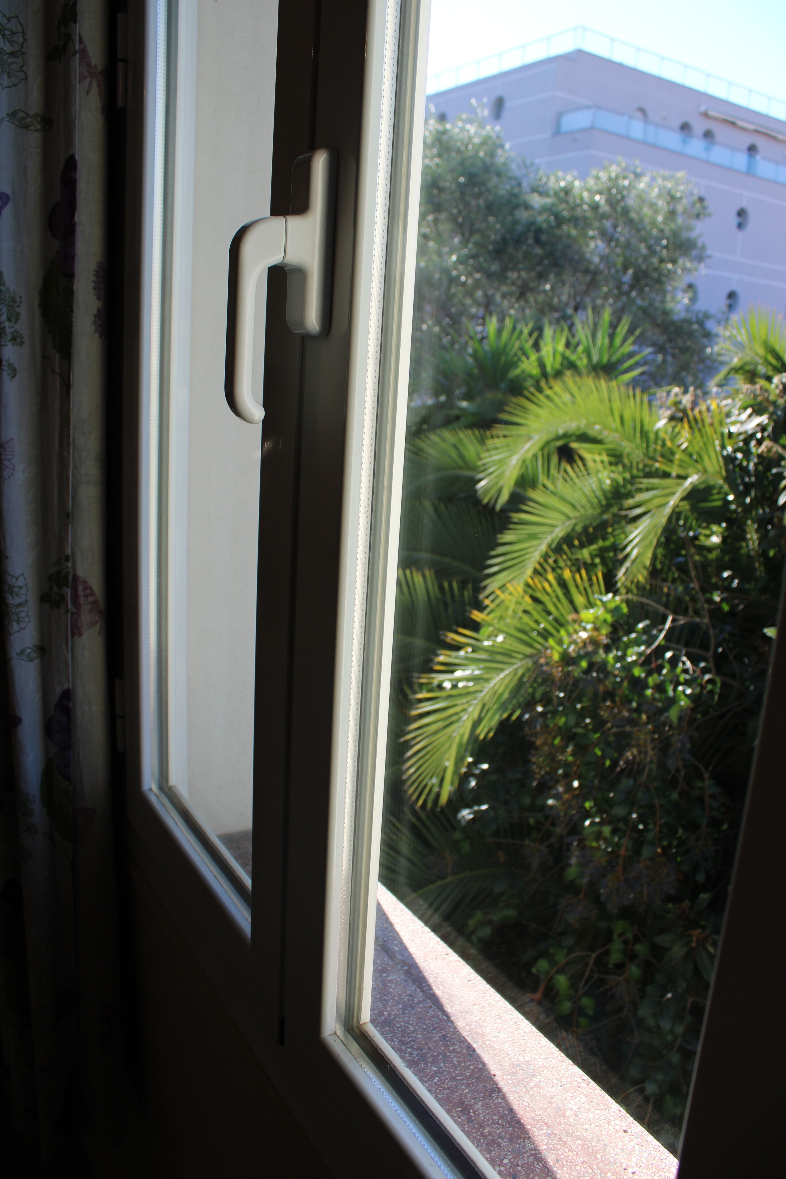 A window with a white frame and a door handle. Outside, there is a building and some greenery.
