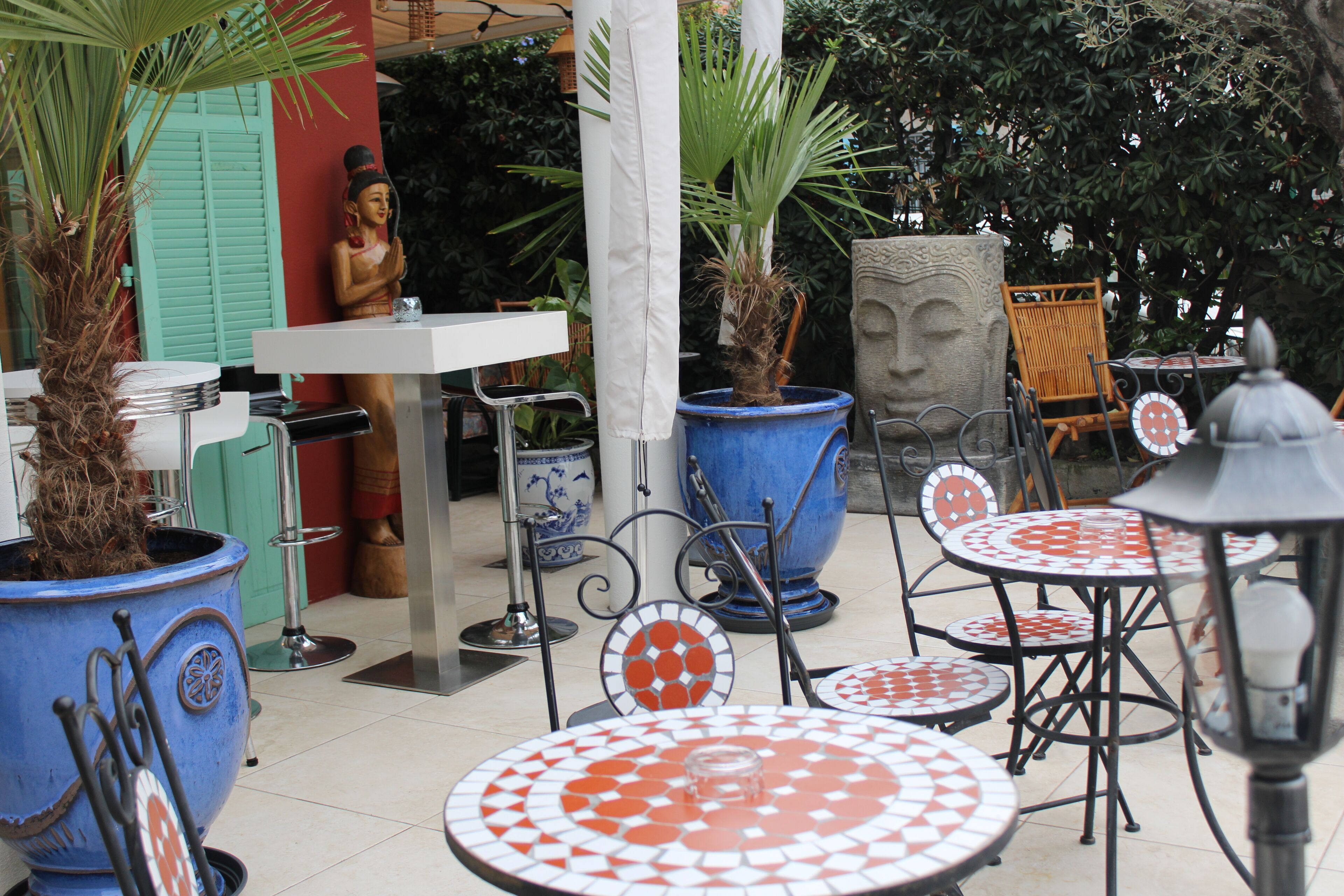 An outdoor seating area with round tables and chairs, large potted plants, and a statue.