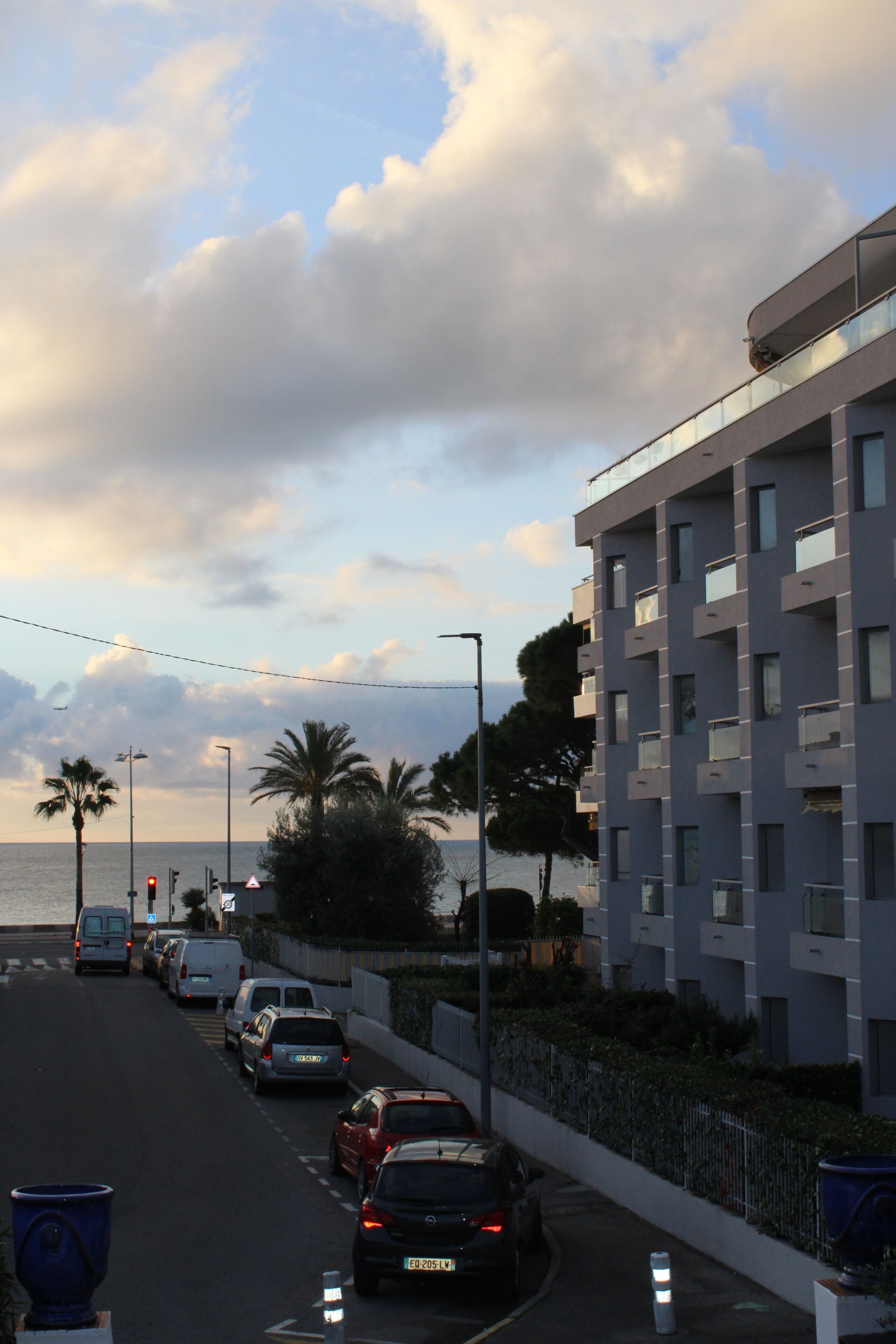 A coastal road with parked cars, a modern building with balconies, and a view of the sea.