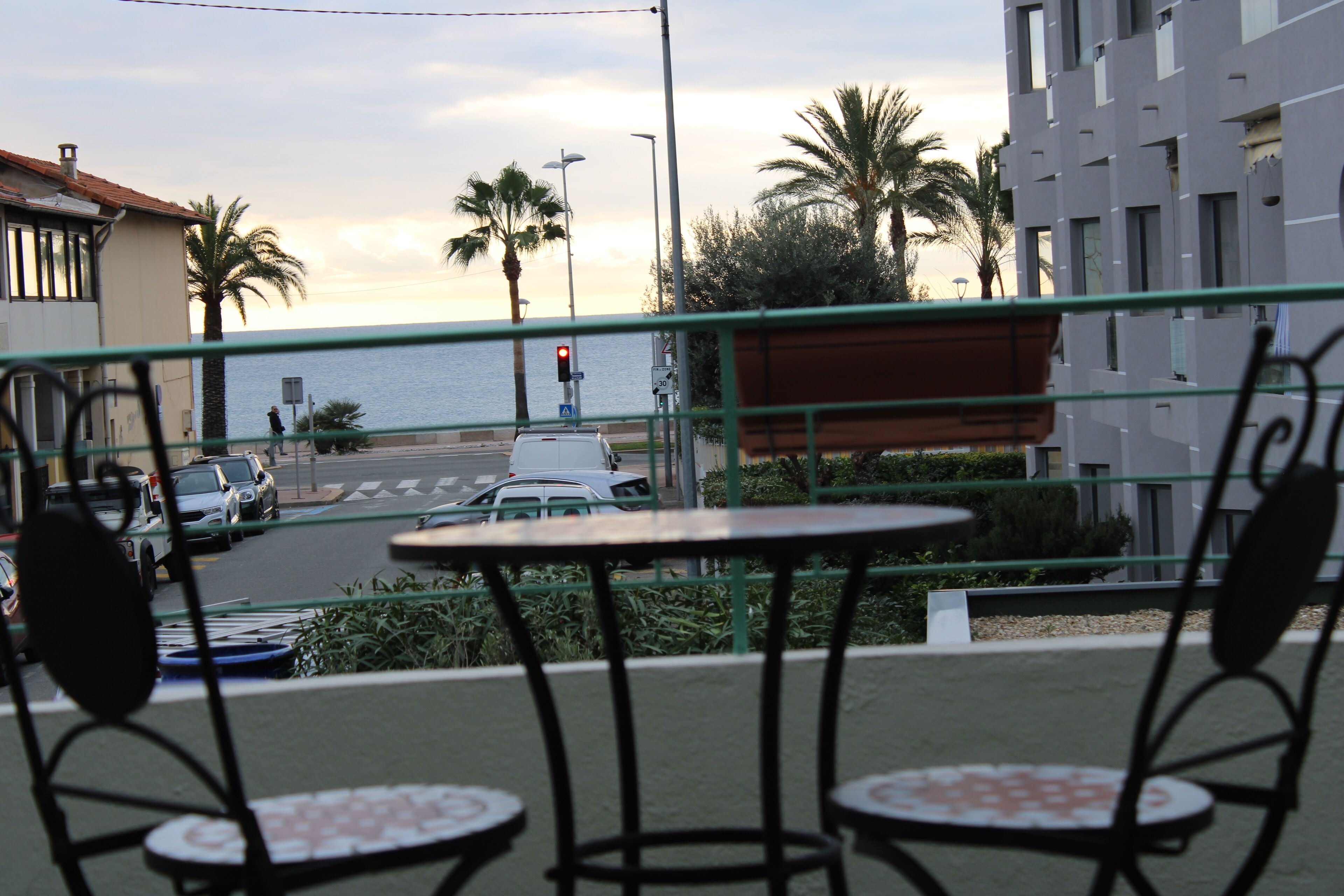 Een balkon met twee tafels en stoelen, met uitzicht op een straat met auto’s en palmbomen, en een gebouw met een balkonhek.