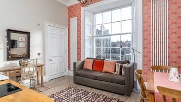 Een gezellige woonkamer met een groot raam, een bank met kussens, een houten tafel en een kamerkleed met een patroon.