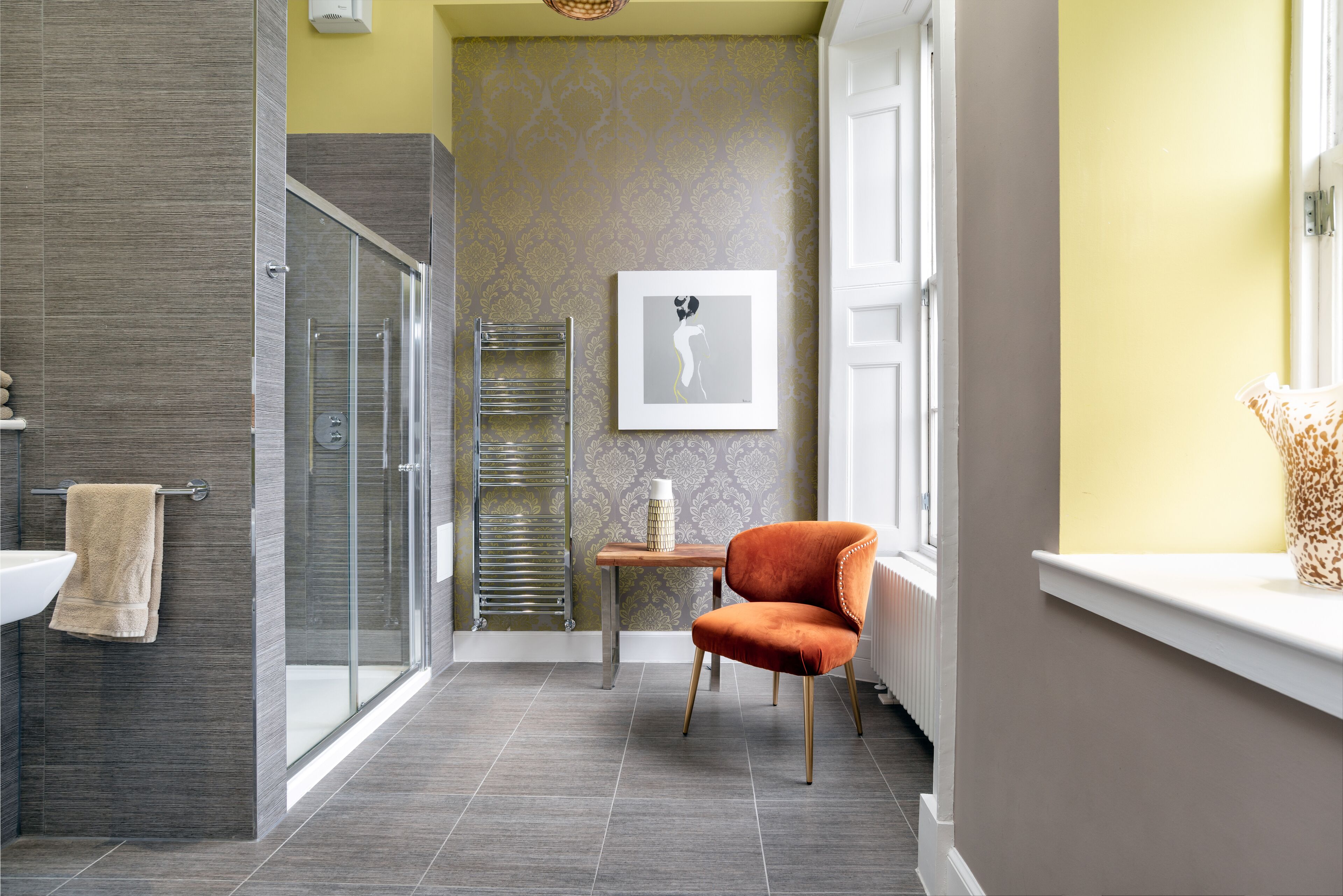 A modern bathroom with a glass shower, a towel rack, a sink, and a chair.
