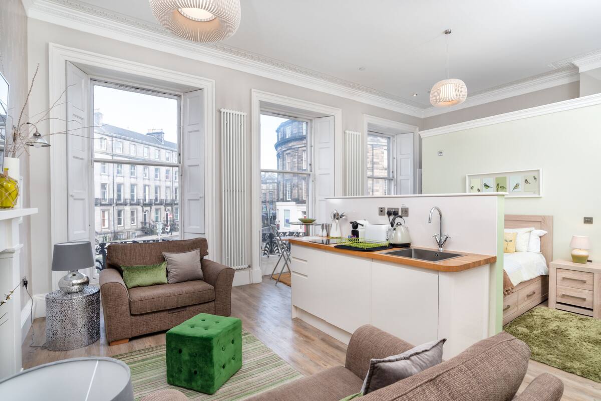 A modern apartment with a kitchen, dining area, and living room. It features large windows, a green ottoman, and a view of the city.