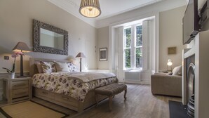 Een slaapkamer met een groot bed, een bankje, een kaptafel, een nachtkastje, een lamp, een spiegel, een raam en een open haard.