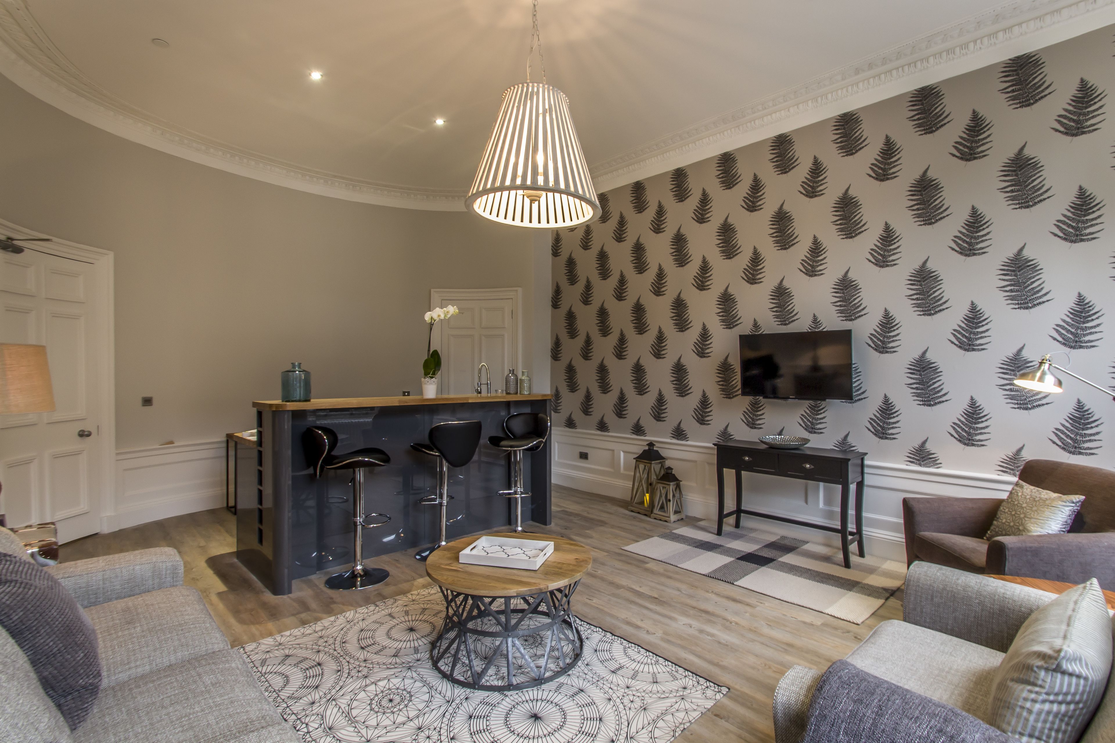 A modern living room with a round coffee table, grey sofas, a patterned rug, a bar area with stools, and a television on the wall.