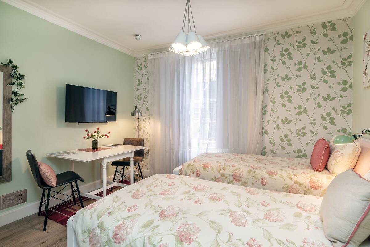 A bedroom with two beds, a desk, a chair, a TV, and a window with curtains.