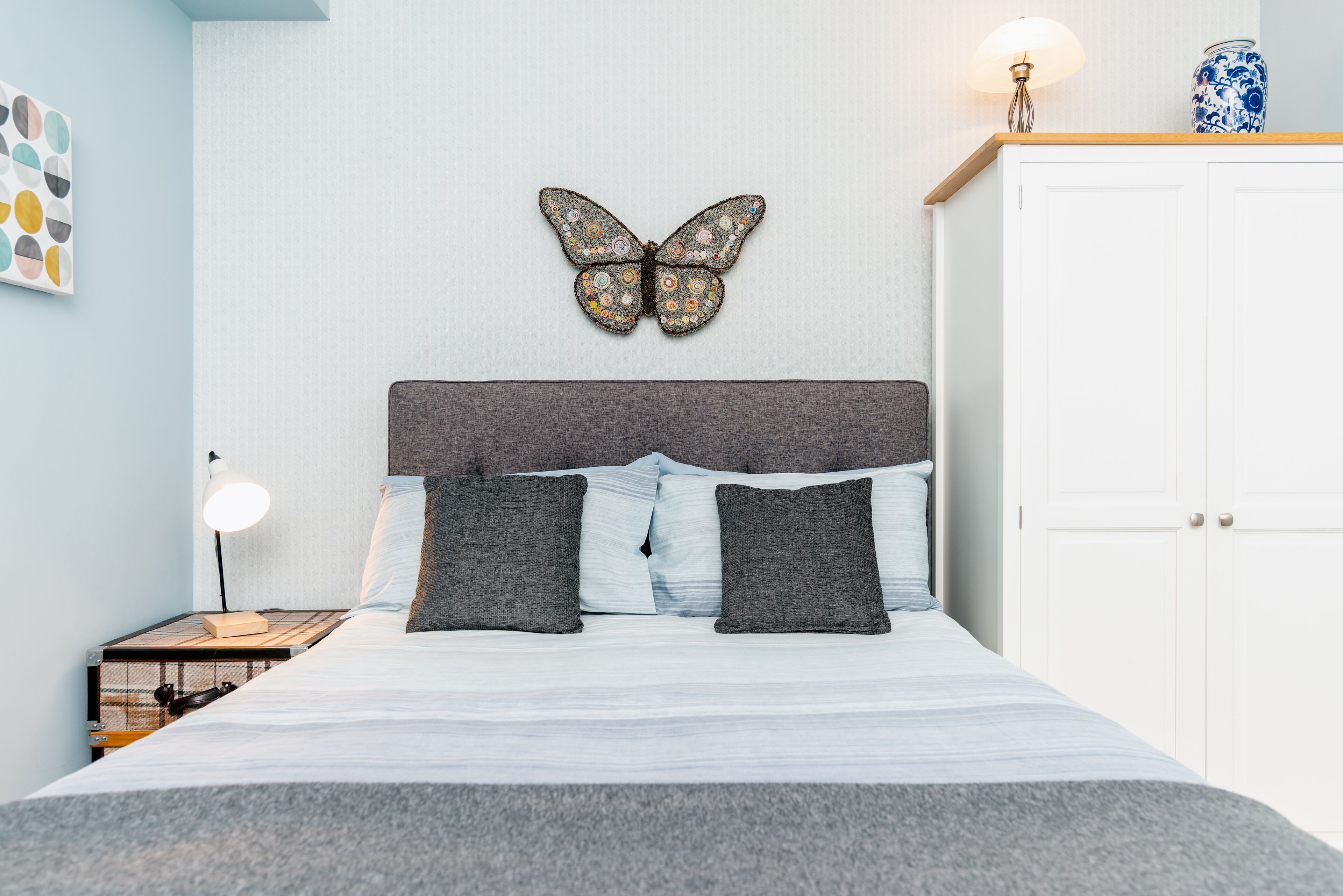 A neatly made bed with a grey headboard, two grey pillows, and a butterfly wall decoration.