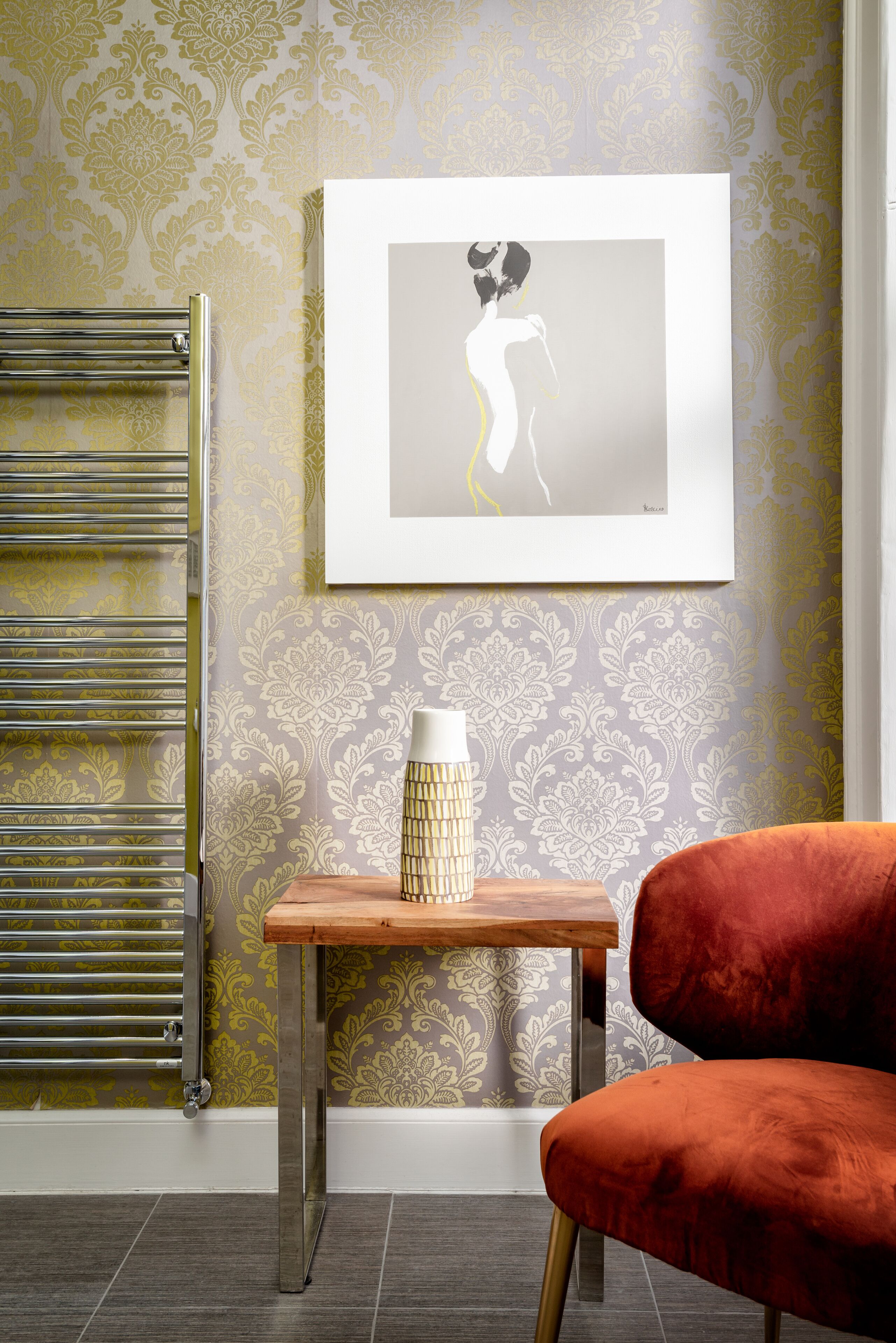 Een kamer met een radiator, een lijst met een kunstwerk aan de muur, een klein houten tafeltje met een vaas, en een rode stoel.