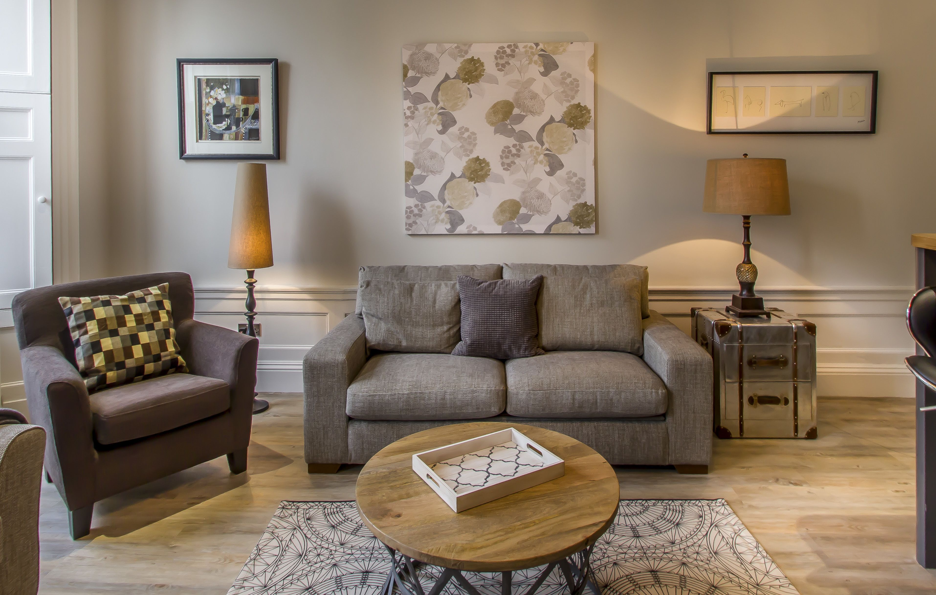 A living room with a sofa, armchair, and a coffee table with a tray. There are two lamps, a rug, and artwork on the wall.