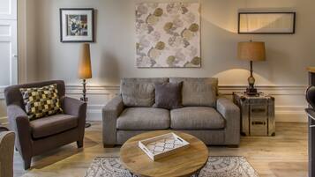 A living room with a sofa, armchair, and a coffee table with a tray. There are two lamps, a rug, and artwork on the wall.