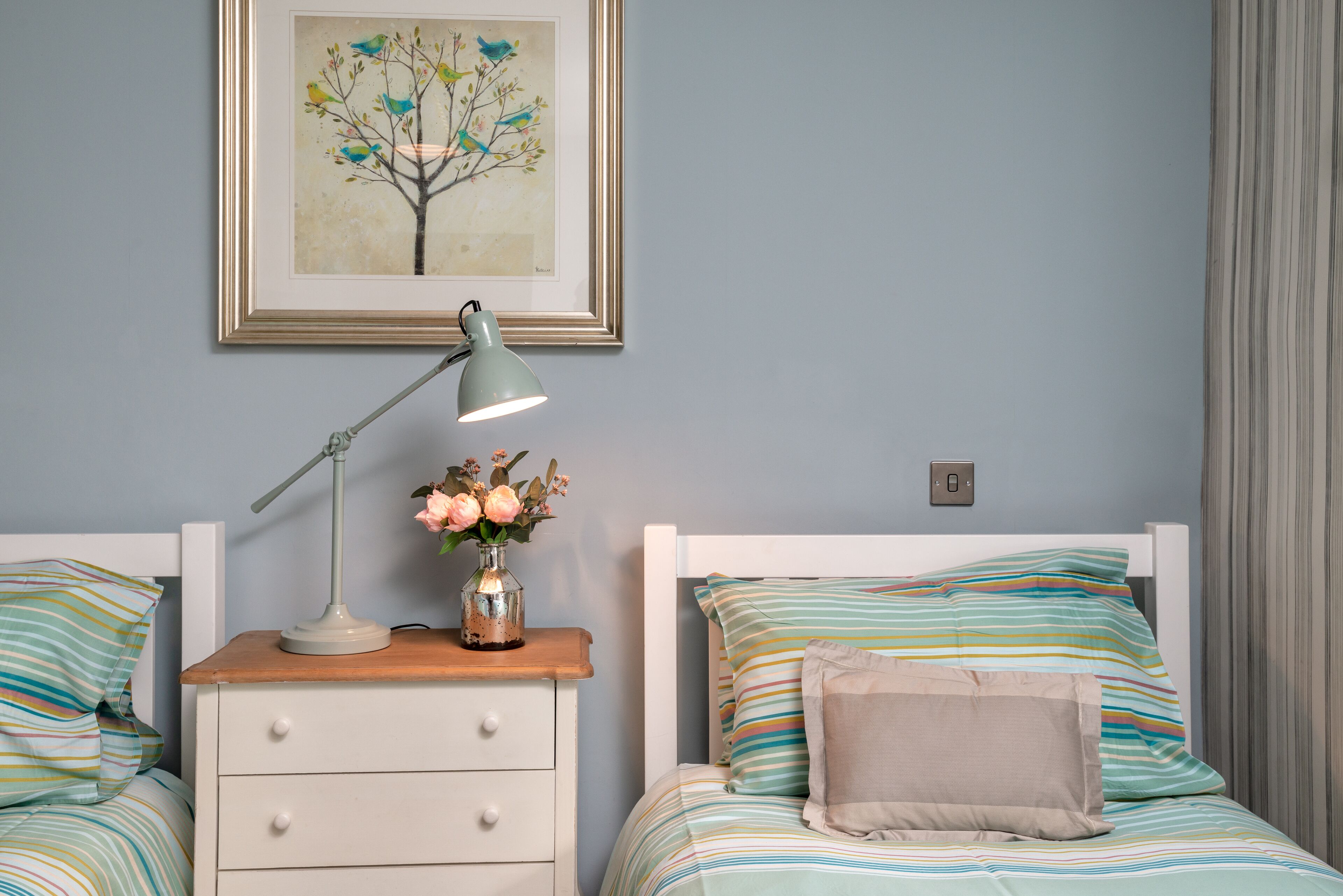 Un dormitorio con mesita de noche, lámpara, un cuadro en la pared y una cama con ropa de cama a rayas.