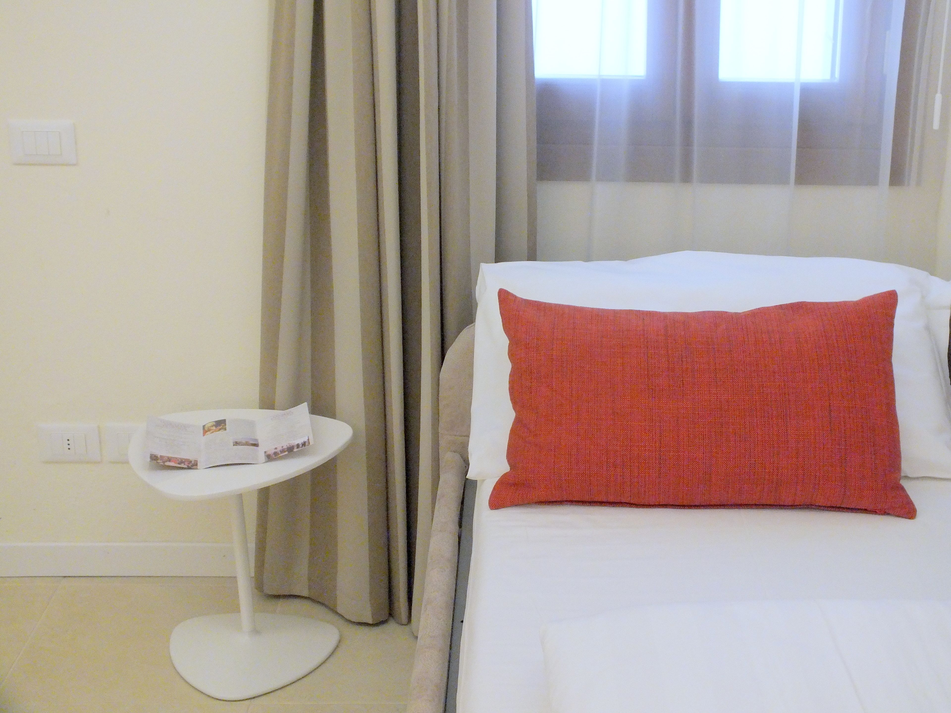 A hotel room with a bed, a small round table with a magazine, and a window with curtains.